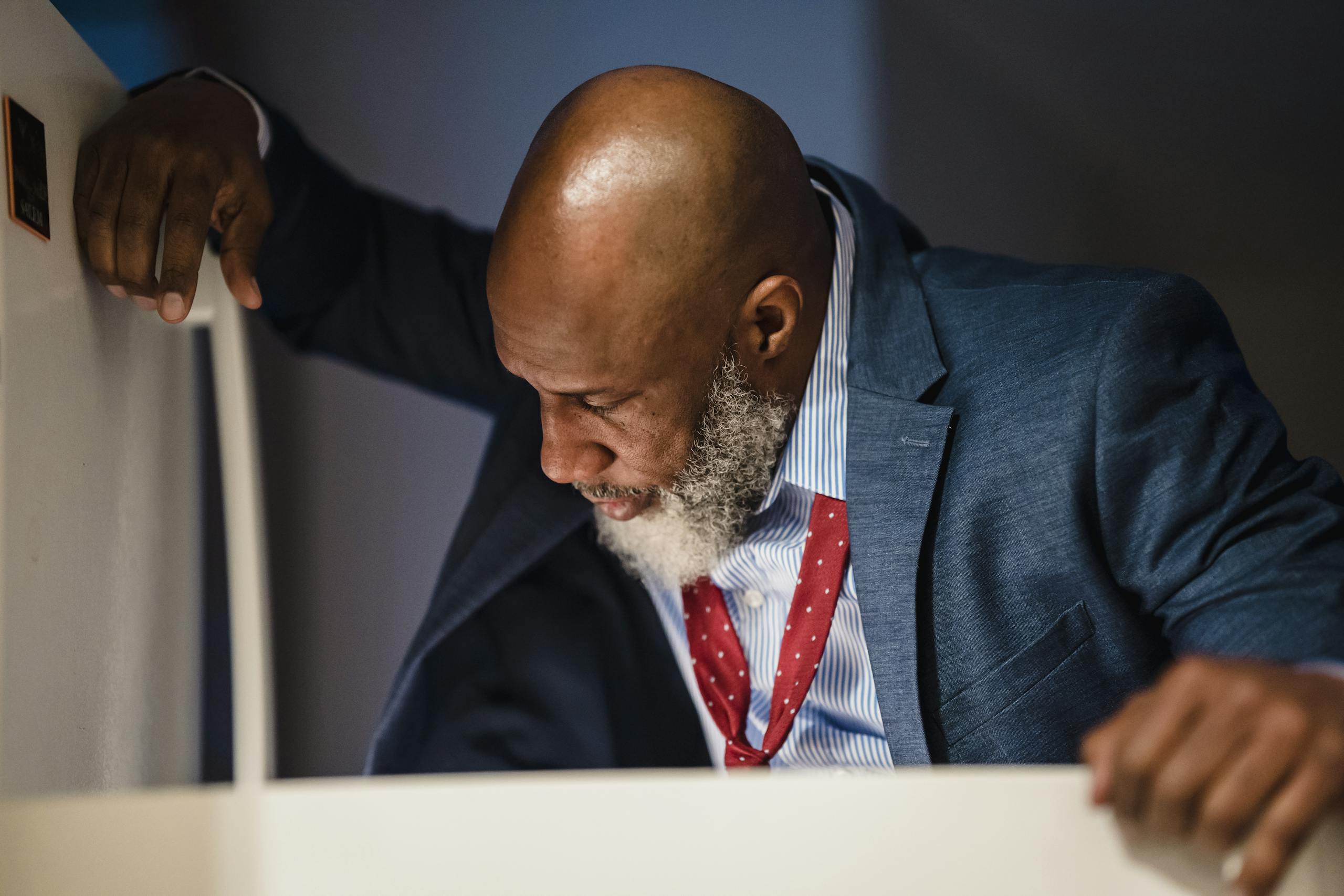 An adult man in a suit looks into an office refrigerator during the daytime.